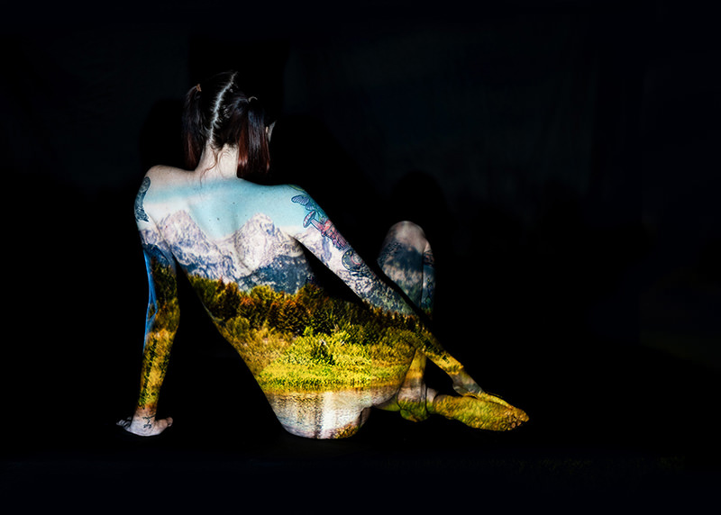 Person sitting against a black background with a detailed mountain, forest, and lake landscape projected onto their back and legs, blending natural scenery with the contours of the body