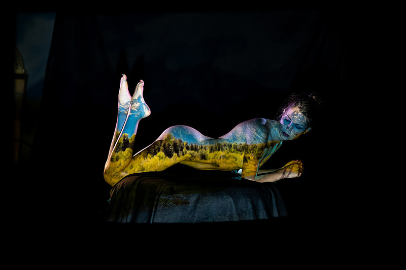 Person lying on stomach with legs raised, nude and body covered in vibrant projection of a landscape featuring trees, green fields, and blue sky, against a dark background