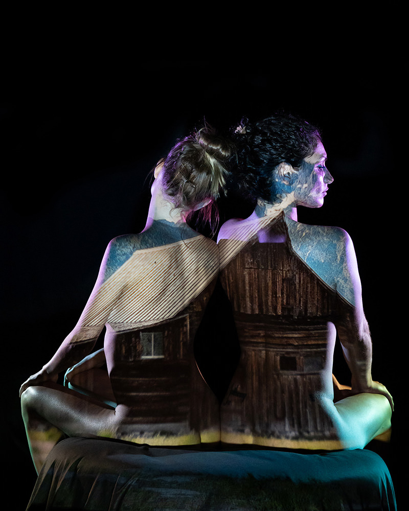 Two women sitting back to back with a projection of a rustic wooden cabin covering their backs and shoulders against a dark background