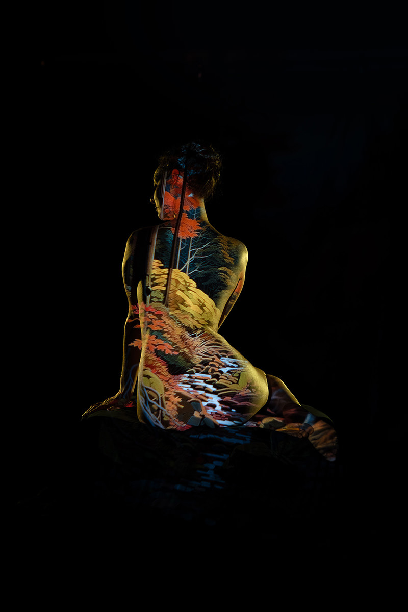 A person sitting with back facing the camera, illuminated by a colorful projection of a forest landscape including trees and a flowing stream, against a dark background