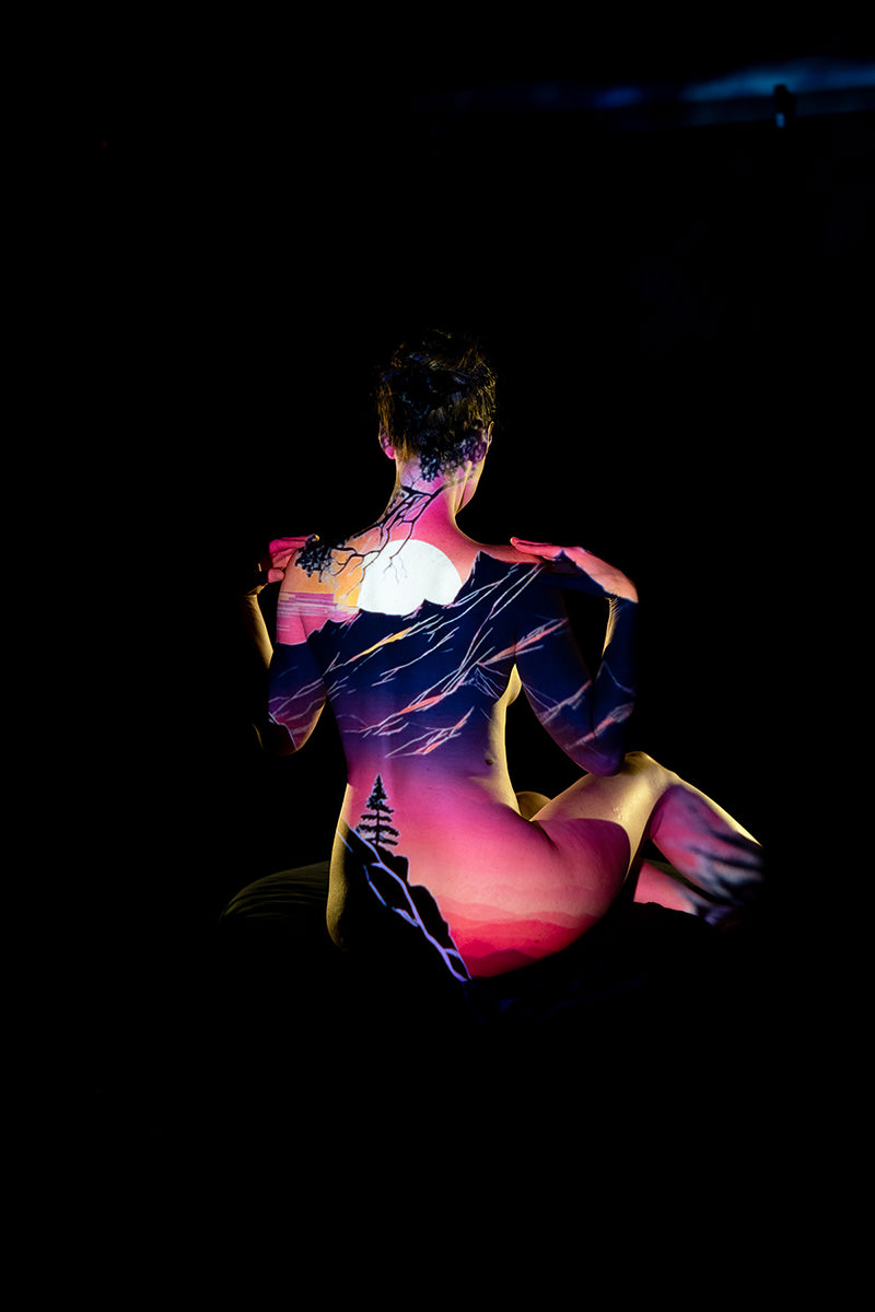 Nude person sitting with their back facing the camera, a vivid sunset mountain landscape projected onto their skin, showing a large sun, tree branches, mountains, and a single pine tree, all against a dark background