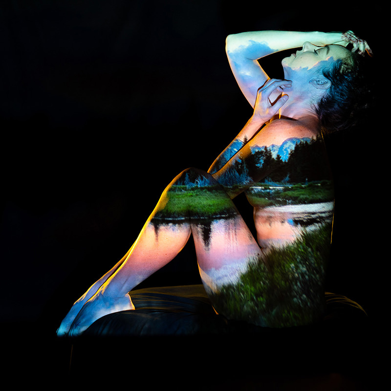 Nude person sitting with knees drawn up and head tilted back, a vibrant landscape of trees, water, and sky projected onto their body against a black background