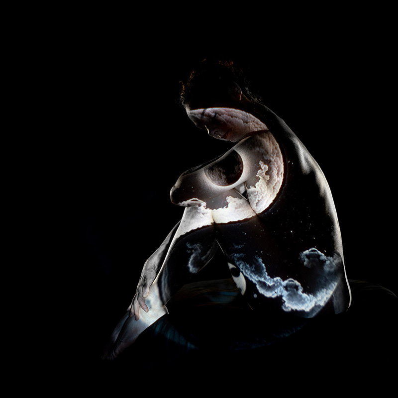 A person seated with knees drawn up, illuminated by a projection of the moon and clouds, against a dark background.