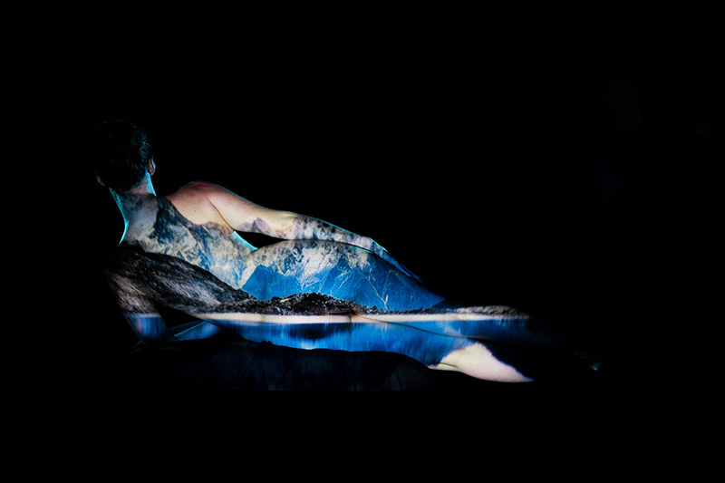 Rear view of a reclining nude person with a mountain landscape projected onto their body against a black background