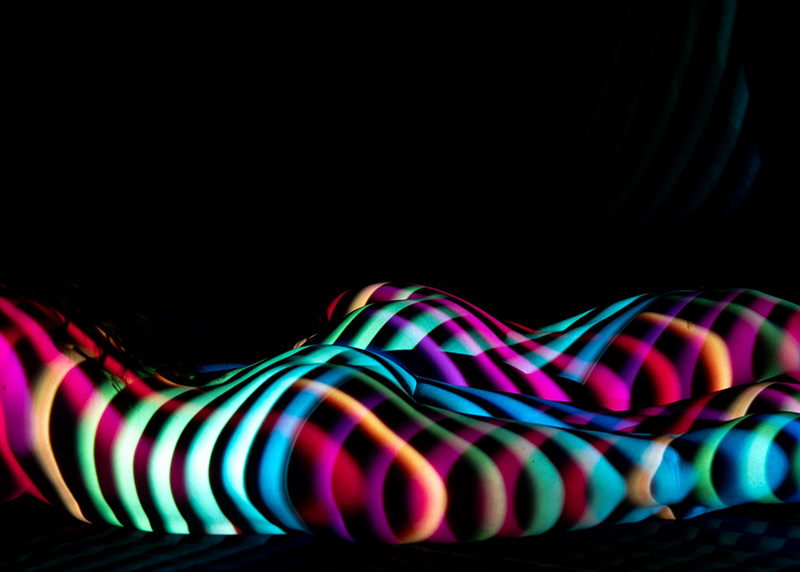 Nude human body lying down with colorful rainbow stripes projected onto the skin against a black background