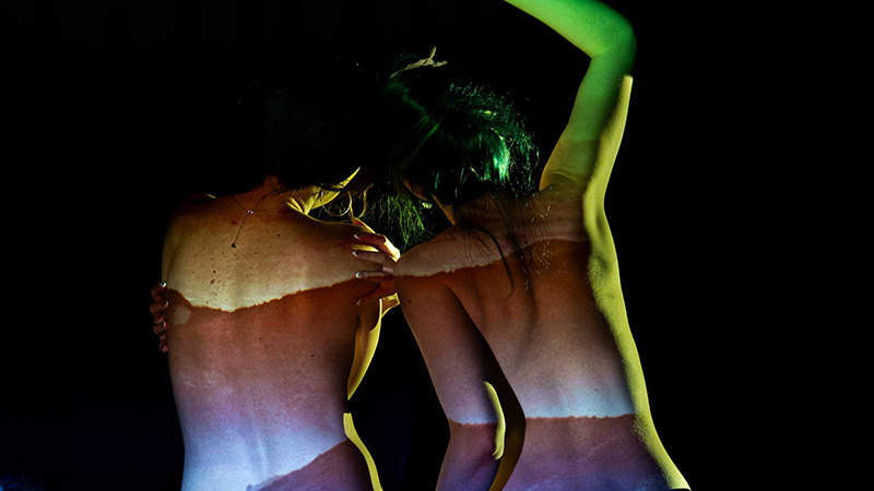 Two people with their backs to the camera, illuminated by colorful light projections creating abstract patterns on their skin against a black background