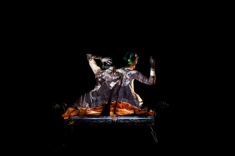 Two nude figures seated on a bench with their backs facing the camera, covered in dark and light earthy patterns projected onto their skin, set against a black background