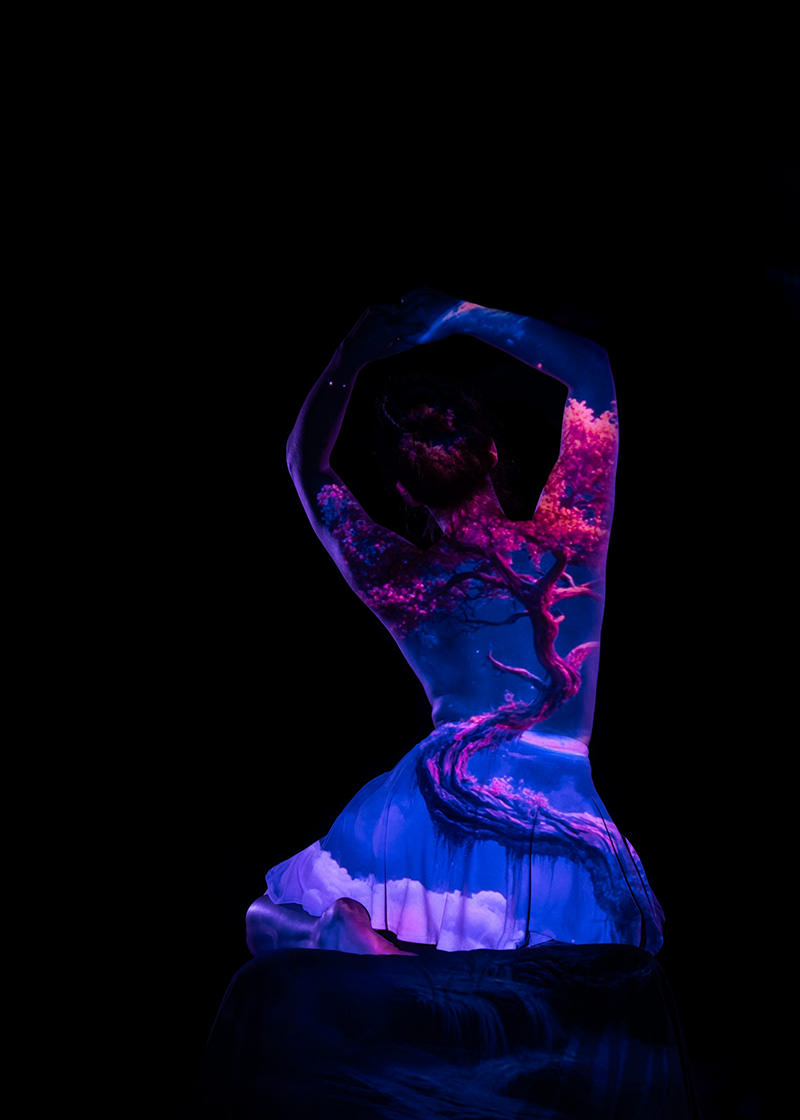 A person sitting with arms raised above their head, with a colorful projection of a twisting tree with pink blossoms and clouds displayed on their back and skirt, set against a black background