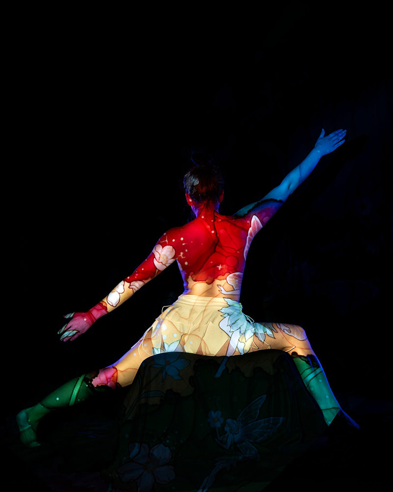 Person sitting with legs extended and arms raised, featuring colorful floral and abstract pattern projections covering their back and legs against a black background