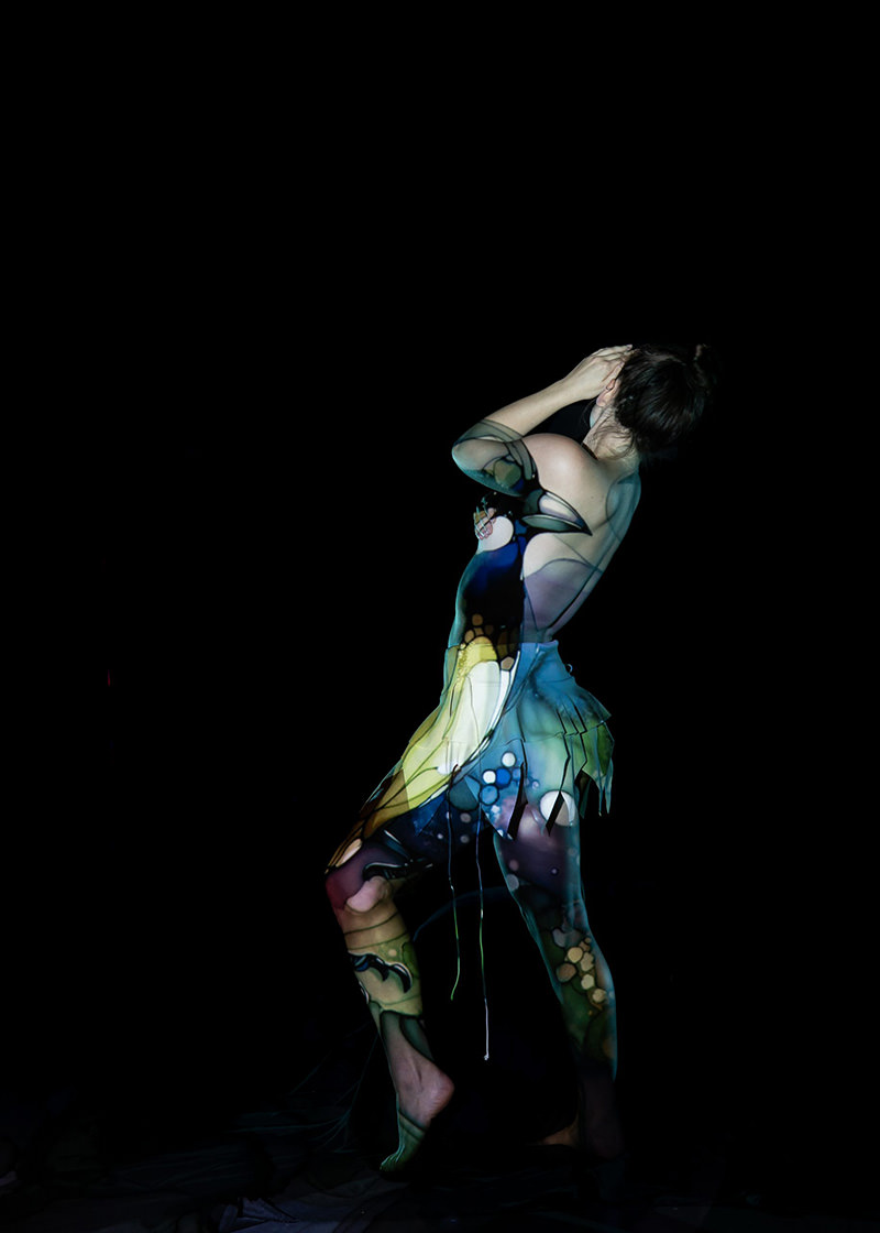 Person posed sideways against black background, colorful abstract patterns projected on their body creating a vibrant and artistic effect
