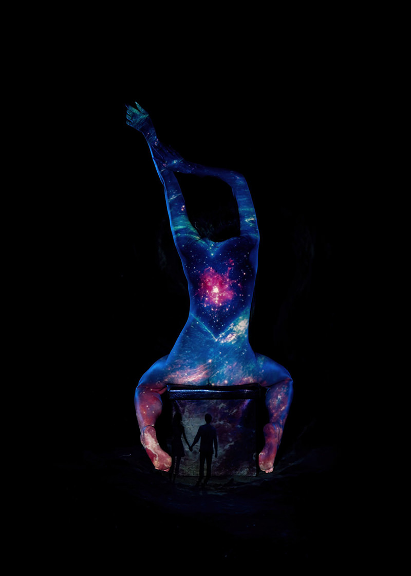 Nude person sitting on a block with arms stretched upward, their back and limbs illuminated by a vibrant galaxy projection, while the block shows a silhouette of a couple holding hands against a starry background, all set against a pitch-black backdrop.