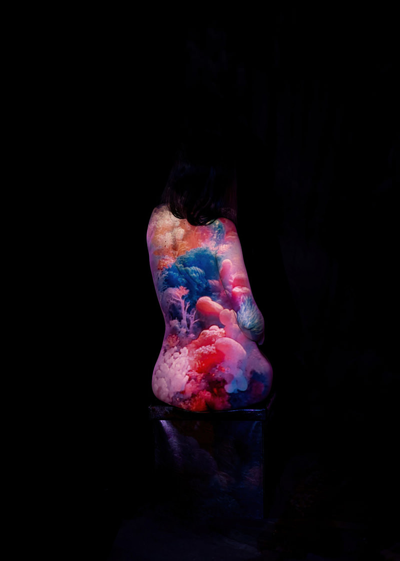 Person sitting with back turned, body painted with vibrant coral reef and underwater sea life patterns in pink, blue, and purple hues against a black background