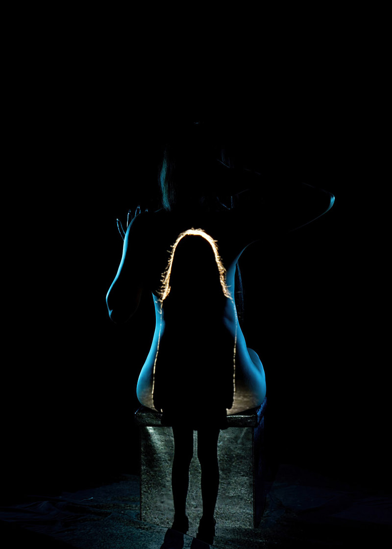 A glowing silhouette of a child's figure is superimposed on and aligned with the back of a seated adult, both surrounded by darkness with subtle blue highlights along the adult's outline.