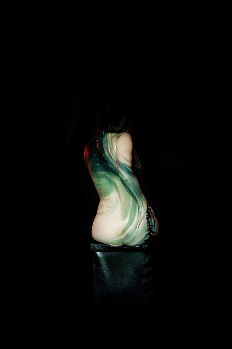 Nude person sitting on a dark stool, back facing the camera, with green and black abstract patterns projected onto their skin, surrounded by a black background