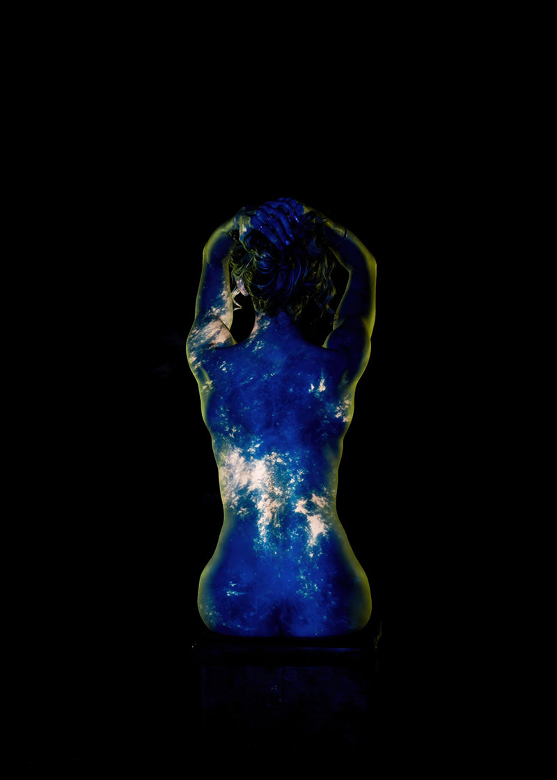 Nude person seen from the back with a projection of Earth’s blue oceans and white clouds covering their skin, set against a black background. The person has their arms raised, hands clasped behind their head.