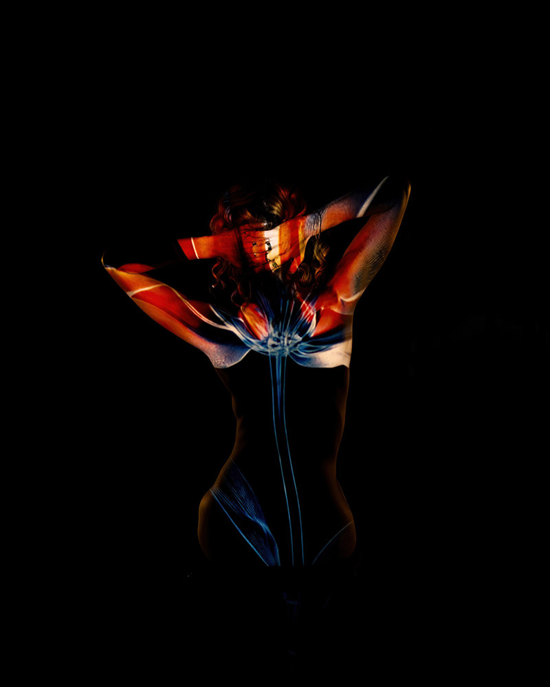 Person standing with back facing camera, arms raised and hands covering head, illuminated by colorful floral light projections against a black background