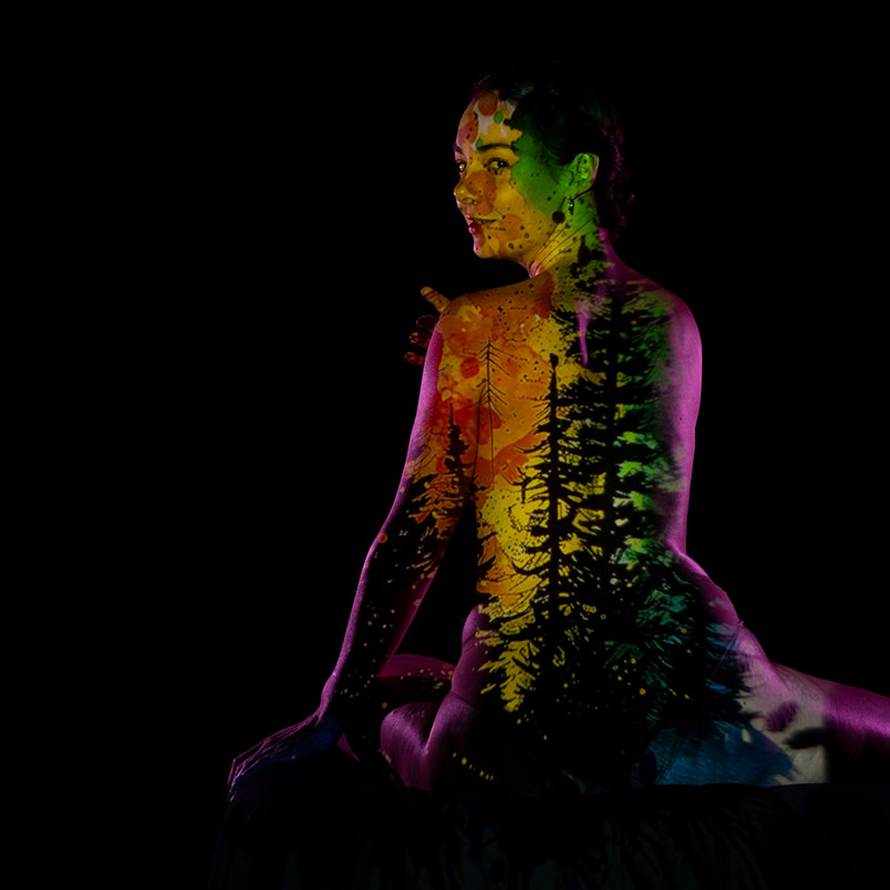 A woman with colorful projections of trees and abstract patterns on her back, sitting against a black background, looking over her shoulder.