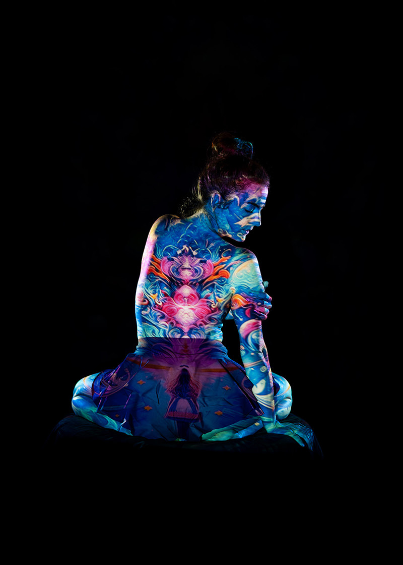 Seated woman with hair in a bun, facing sideways with vibrant and colorful body art covering her back, arm, face, and skirt, against a black background