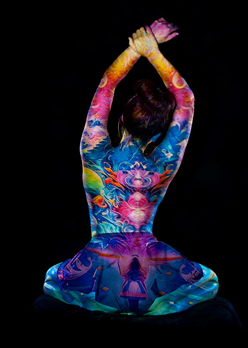Person seated with back facing viewer, arms crossed above head, covered in a vibrant, multicolored projection of swirling patterns and a small figure of a girl on a swing around the waist, against a black background