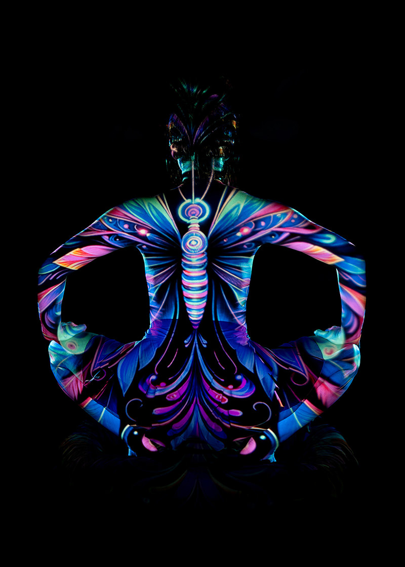 Person seated cross-legged facing away with meditative pose, covered in vivid neon butterfly body paint patterns against a black background