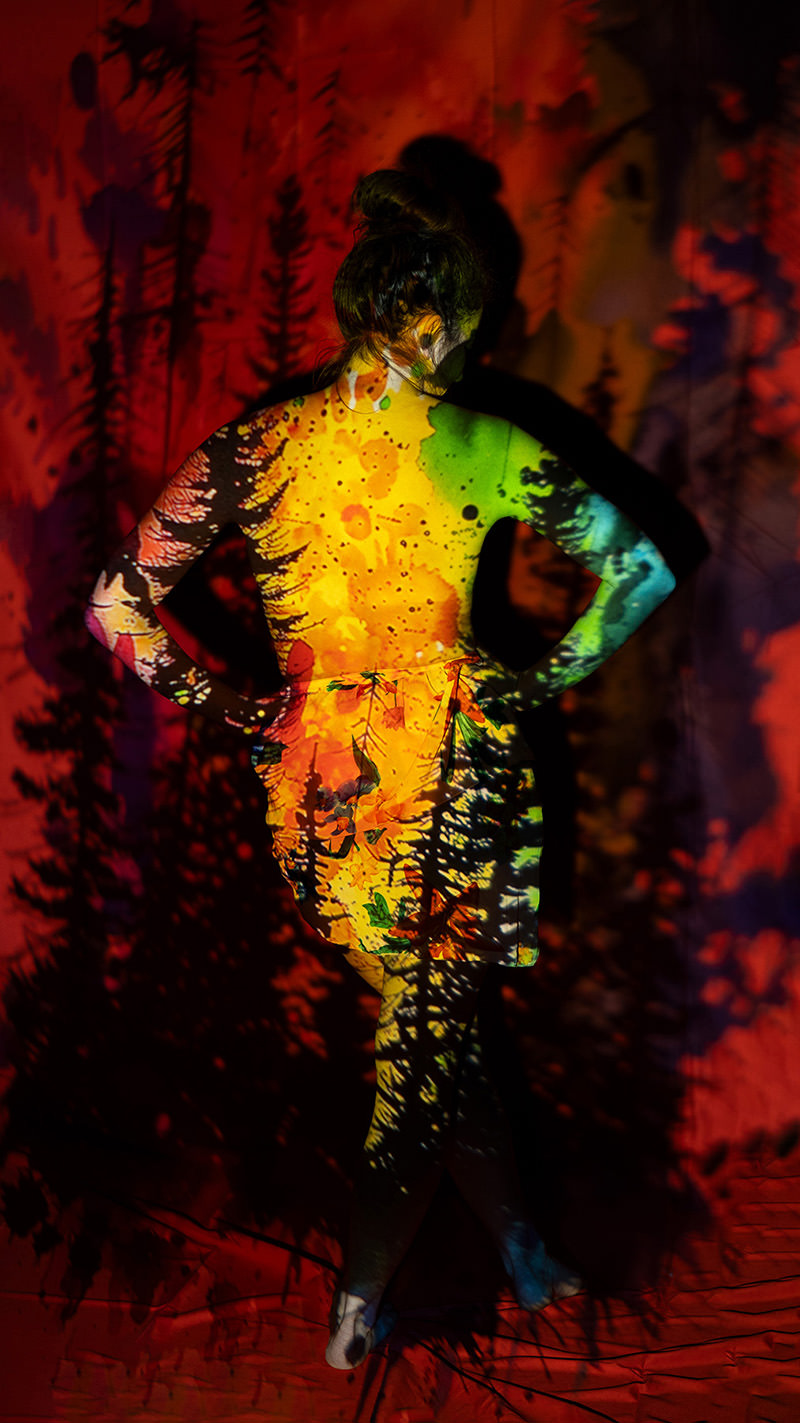 Person standing with arms akimbo, back facing camera, covered in vibrant multicolored nature-inspired projections on their skin and background