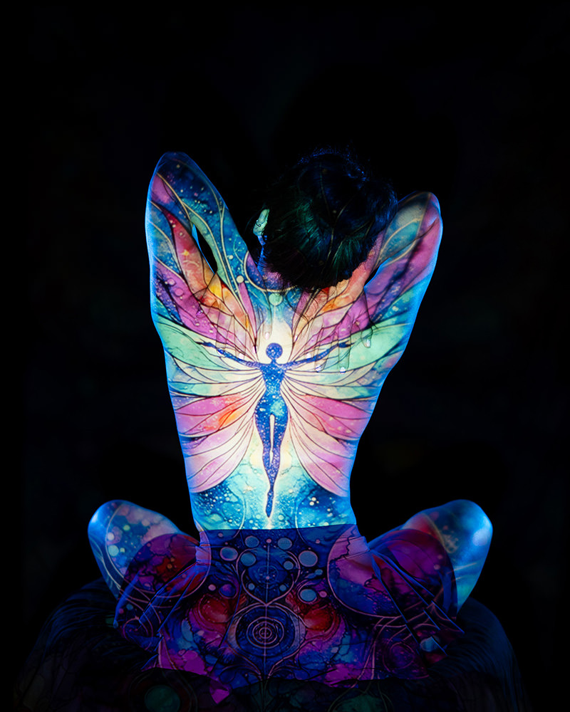 Person sitting with back facing camera, illuminated with colorful projection of a fairy with large, intricate wings covering the entire back, set against a dark background.
