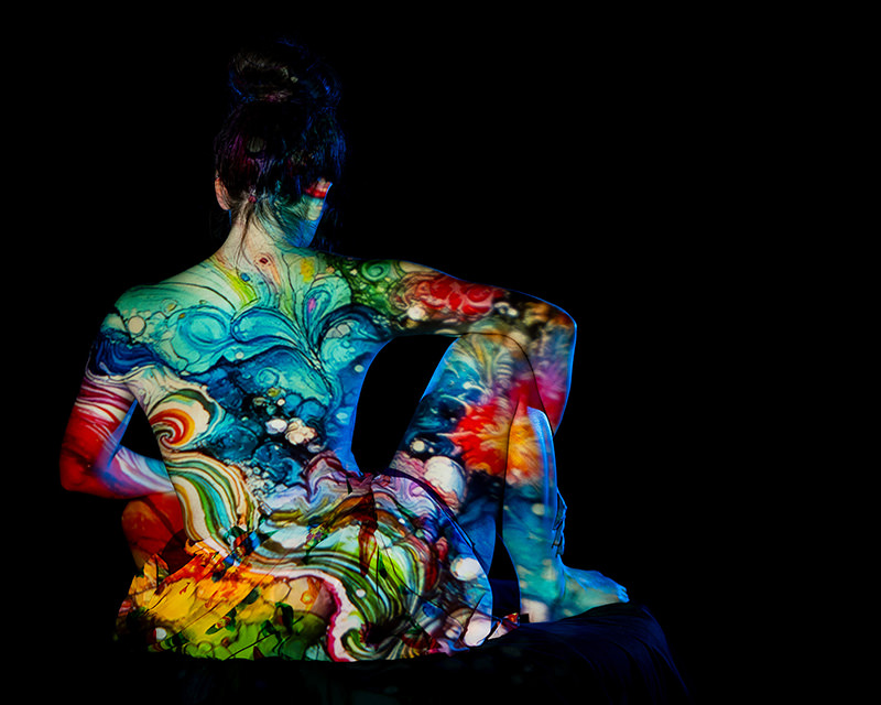 Back view of a seated person with dark hair tied up, covered in swirling, colorful abstract light projections against a black background