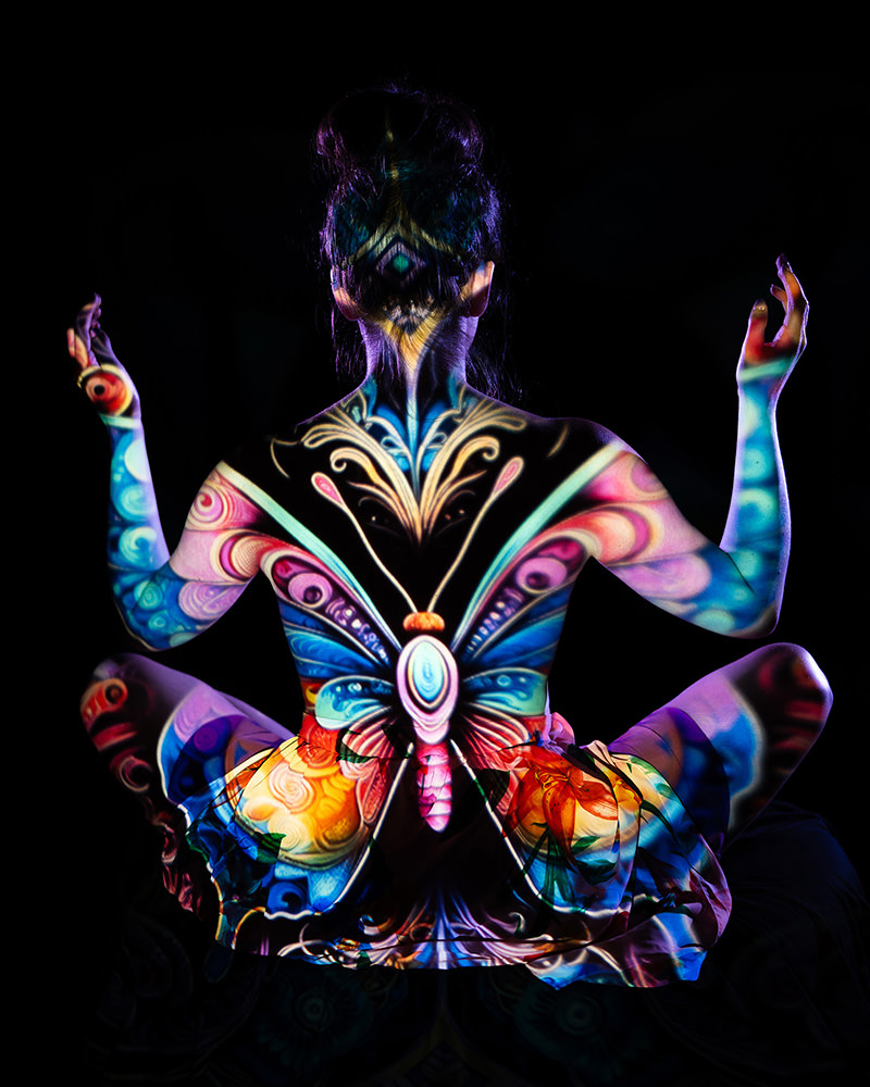 Person sitting cross-legged facing away with colorful butterfly and abstract designs projected onto their back, arms, and legs against a black background