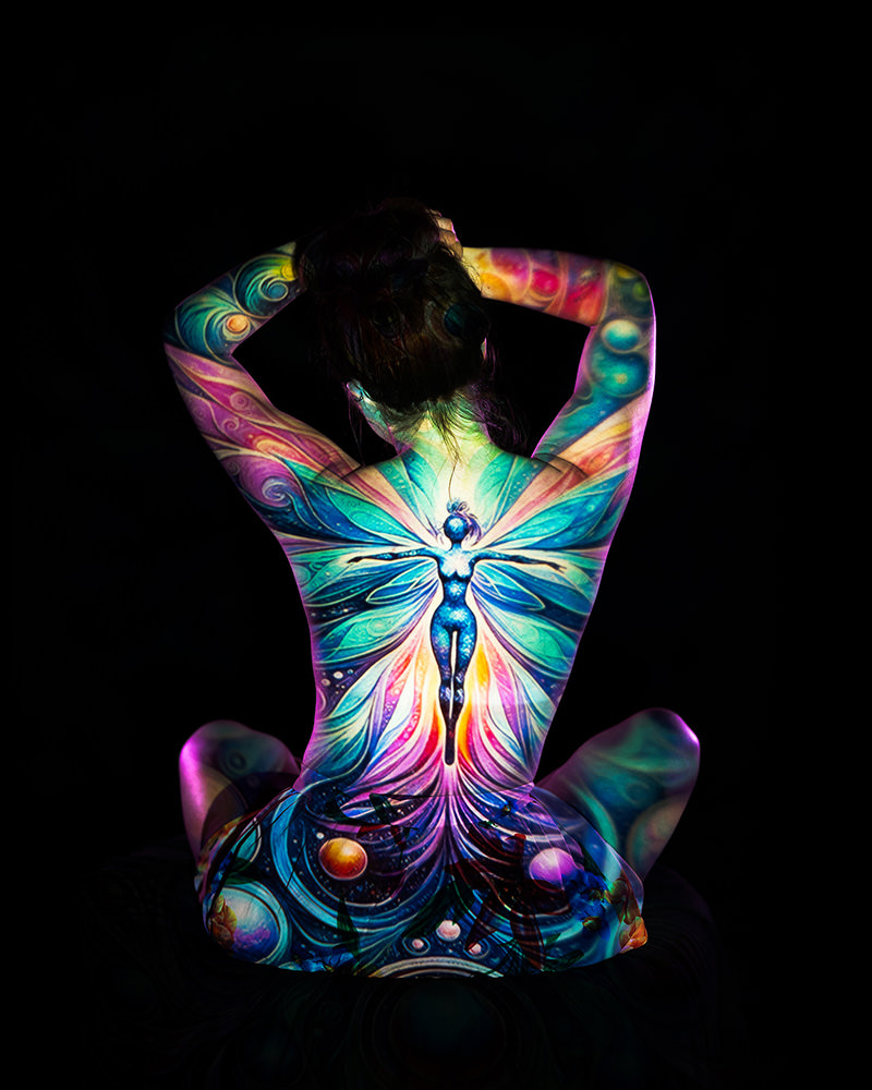 Person seated cross-legged facing away, with colorful, glowing body art depicting a radiant fairy with outstretched wings covering the entire back and extending onto arms and legs against a black background