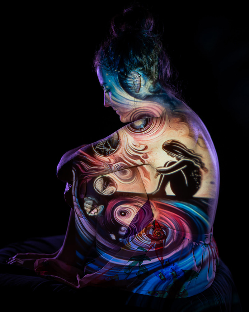 A person sitting in a dark space with colorful, surreal art projections cast across their bare back and arms, featuring swirls, butterflies, and a silhouette of a seated figure