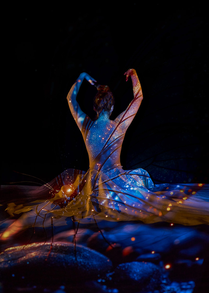 Person with hair in a bun sitting with arms raised, their back illuminated by vibrant blue and orange light projections resembling stars and cosmic patterns against a dark background