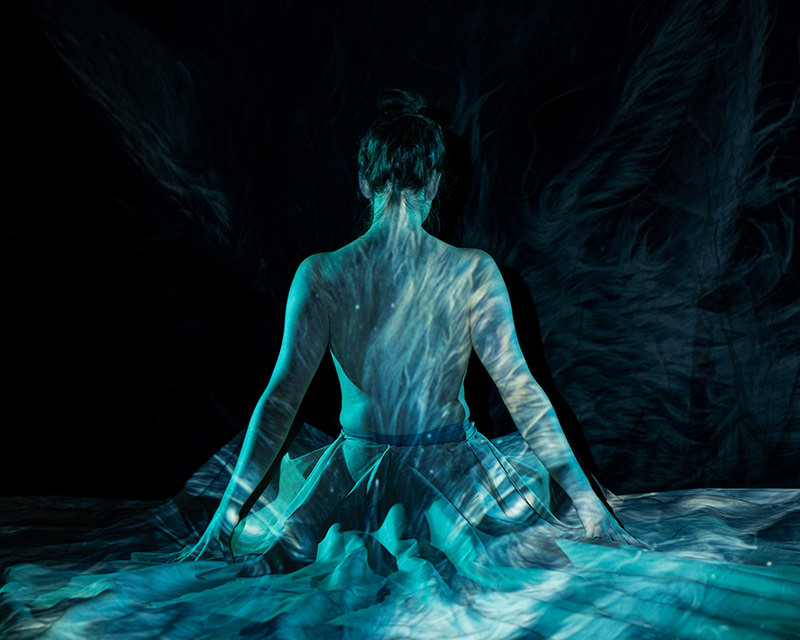 Back of a person sitting with arms resting on the floor, bare upper back exposed, with blue and white swirling light patterns projected onto their skin and surrounding fabric in a dark space