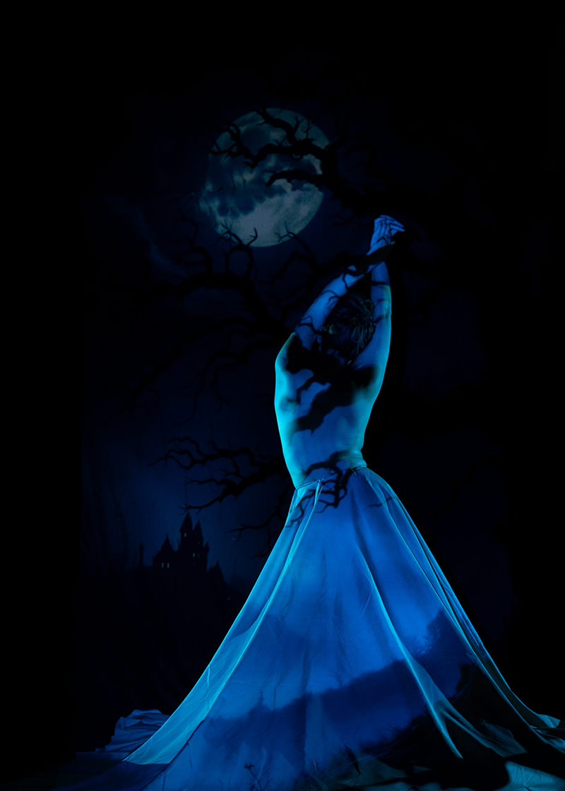 A person with a bare back stands with arms raised, draped in a flowing blue fabric. Shadows of twisted tree branches fall over their body, set against a large full moon and dark night sky.