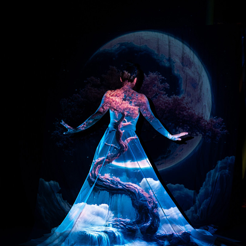 Back view of a person standing with arms outstretched, a vivid projection of a twisted tree, clouds, and a large moon covering their body and flowing dress against a dark background.