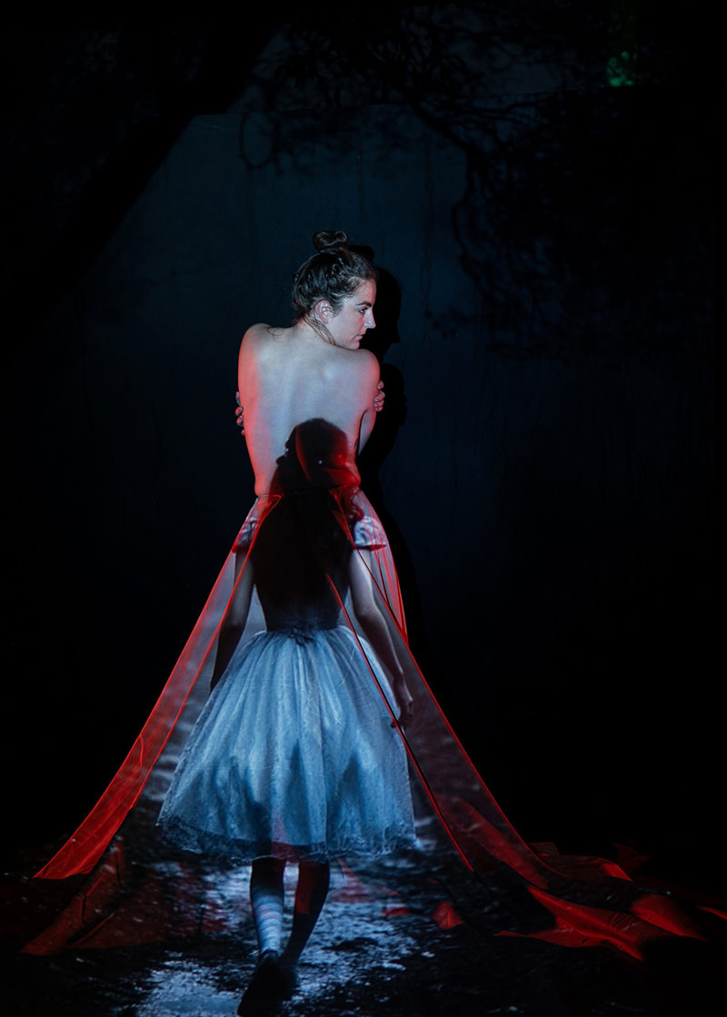 A woman with bare back stands against a dark background, with a translucent overlay of a young girl in a white dress walking away, creating a double exposure effect with a flowing red veil.