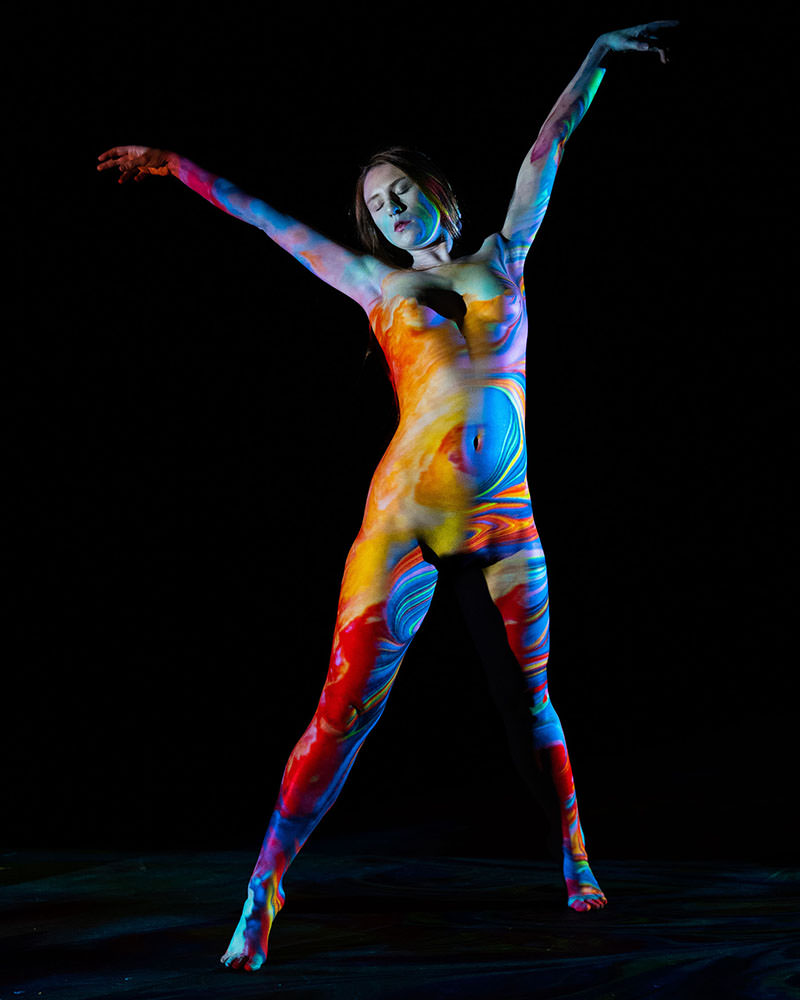 Nude woman with colorful swirling body paint standing with arms extended, eyes closed, against a black background