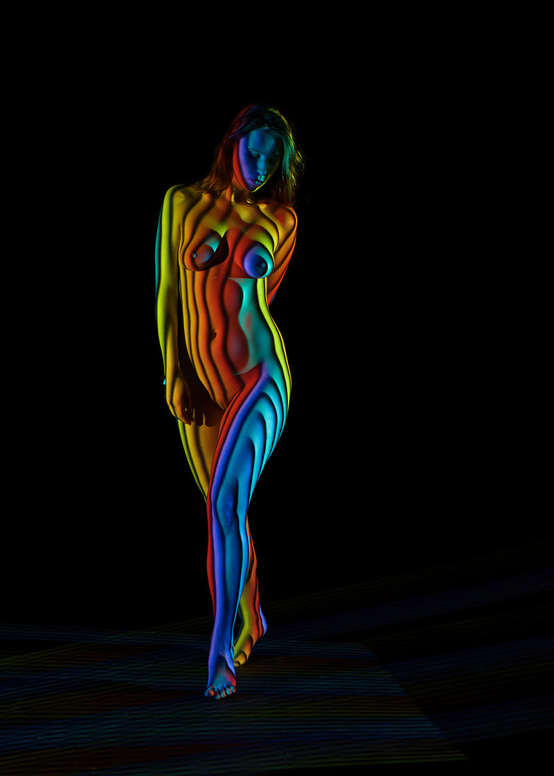 Naked woman standing with her head tilted down and one leg slightly crossed over the other, illuminated by colorful rainbow light patterns creating stripes on her body against a black background