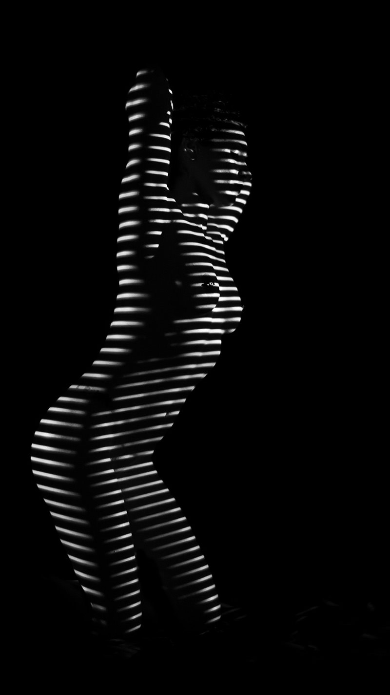 Nude person standing with arms raised, illuminated by horizontal white light stripes creating a zebra-like pattern across the body against a black background