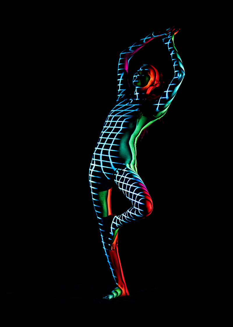 Person striking a balanced pose with arms raised, covered in a blue and white grid pattern projection with red and green lighting effects, against a black background