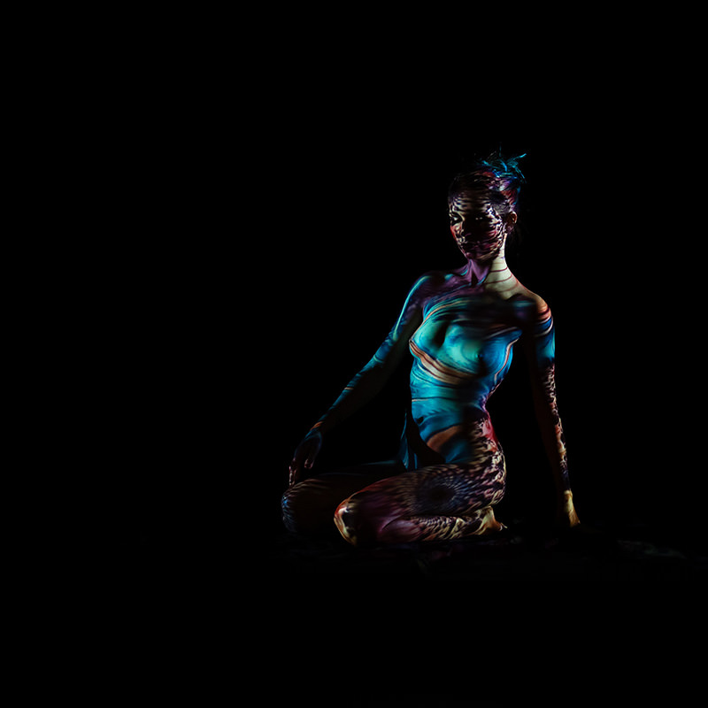 Nude person kneeling with multicolored light patterns projected across their body against a black background