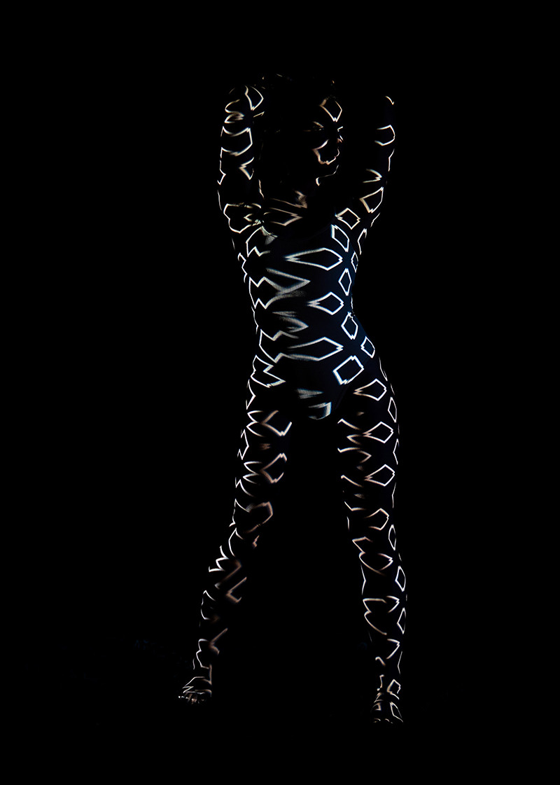 A person standing with arms raised and hands behind head, their body covered in glowing white geometric shapes against a black background, creating a striking silhouette effect.