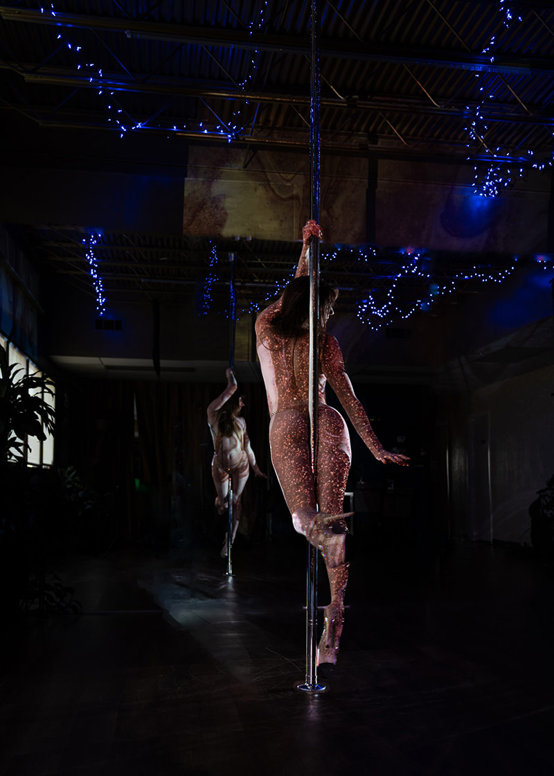 A woman performing pole dance moves in a dark studio with blue string lights on the ceiling, her body covered in small glowing dots, and a large mirror reflecting her image.