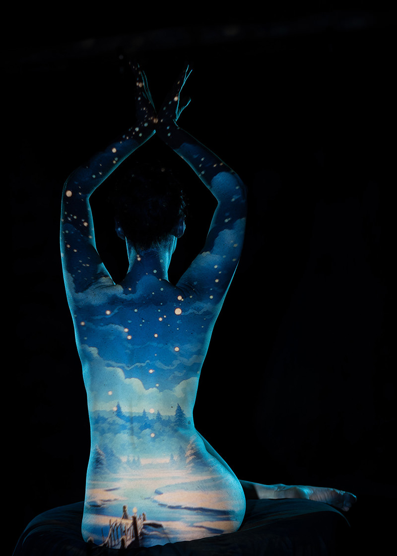 Nude person sitting with back facing the viewer, arms raised overhead, while an artistic projection of a snowy winter landscape with trees, a river, and falling snowflakes covers their back