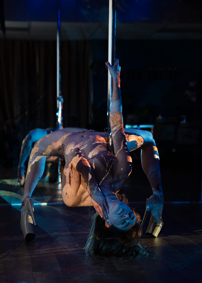 A woman performing an advanced pole dance move, arching her back around the pole with one hand gripping it and the other touching her face, wearing high heels and minimal clothing. Blue and orange light patterns are projected onto her body and the dark room floor.
