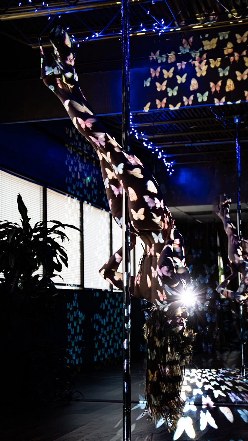 Person performing an upside-down pole dance move with colorful butterfly light projections covering their body and surroundings, including a mirror reflecting the scene, in a dimly lit room with window blinds and blue string lights.