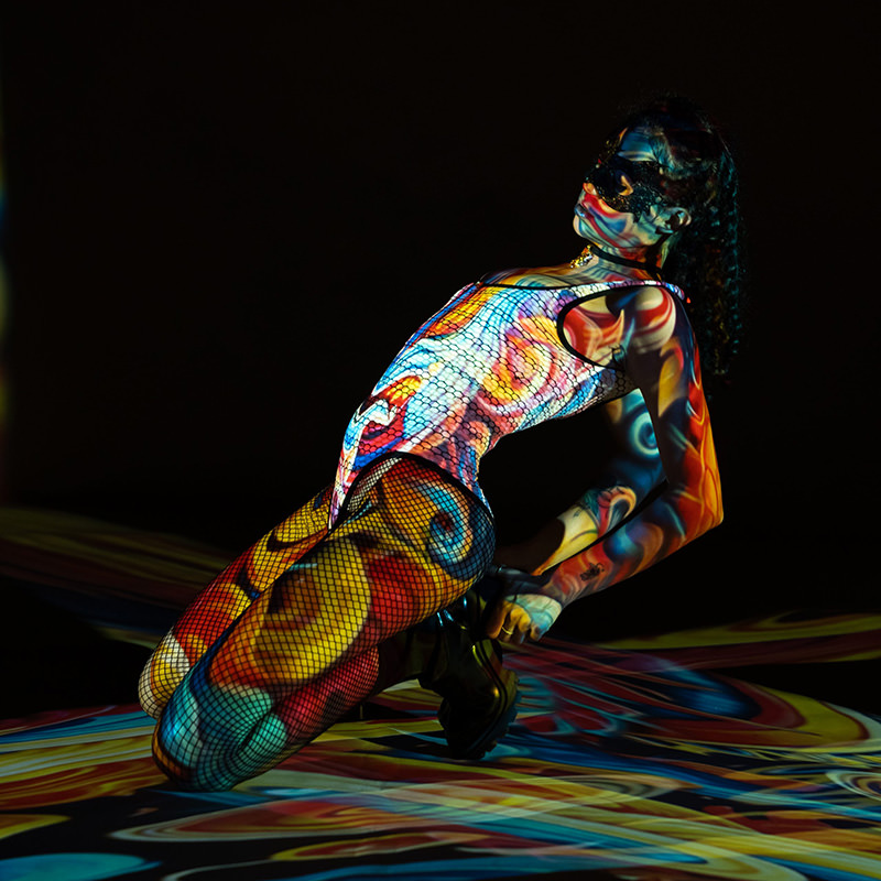 Person leaning back on knees with colorful abstract patterns projected on body, wearing fishnet tights, high boots, and a black mask, against a dark background