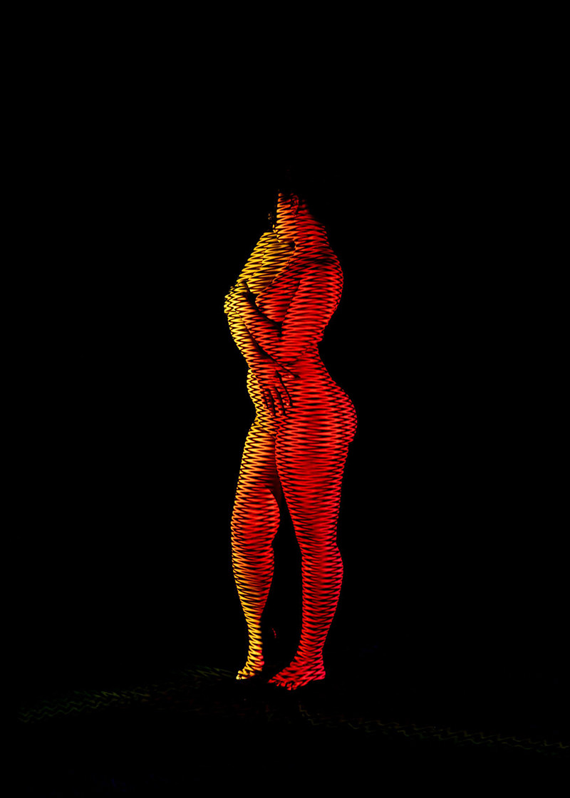 Nude standing person with arms crossed, illuminated by red and yellow zigzag light projections against a black background