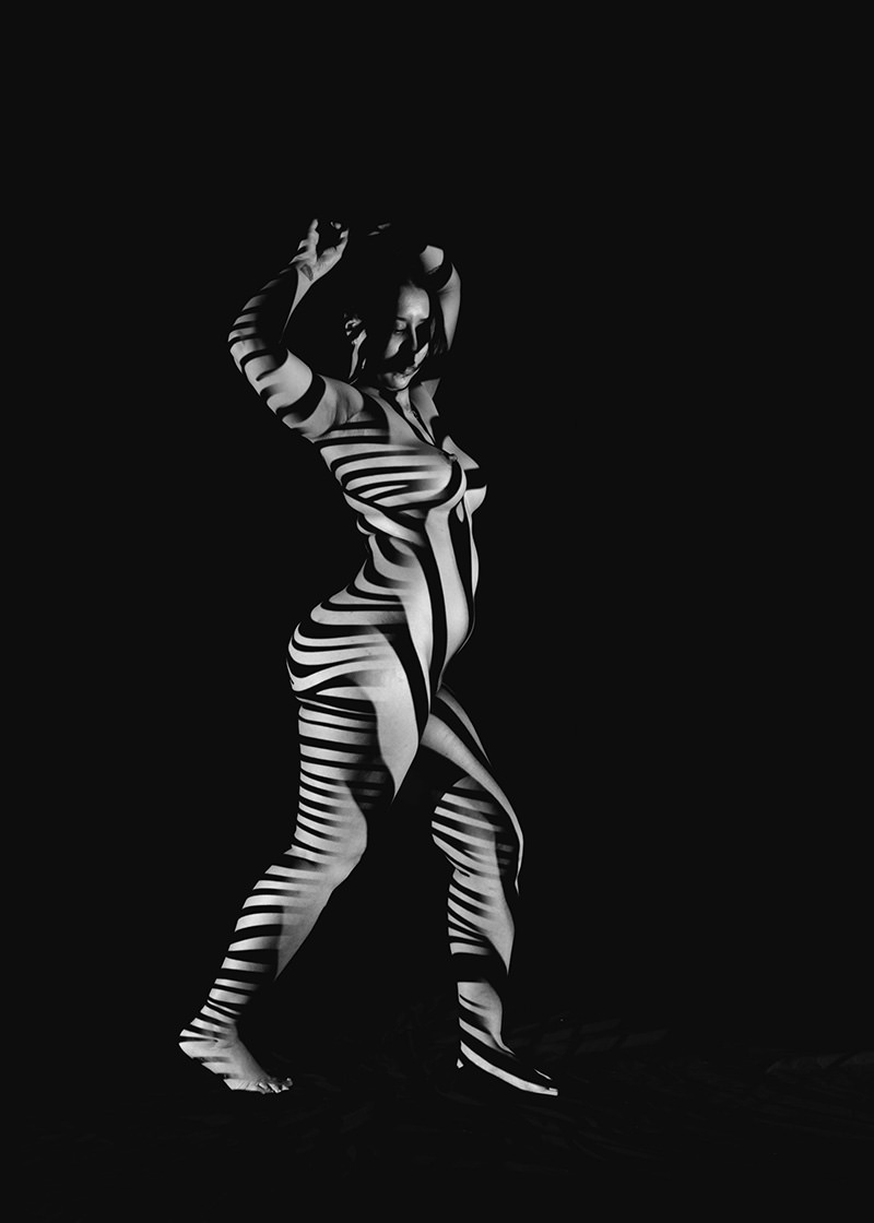 Nude person standing with arms raised, covered in black and white striped shadows against a dark background