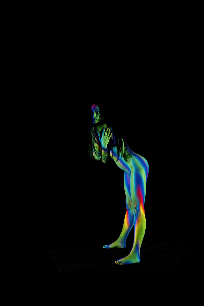 A nude woman posed against a black background, her body illuminated by multicolored light patterns in blue, green, red, and yellow hues highlighting her curves and contours. She is standing on tiptoe, leaning forward slightly with her hands crossed in front of her chest, and long dark hair falling over her shoulders.