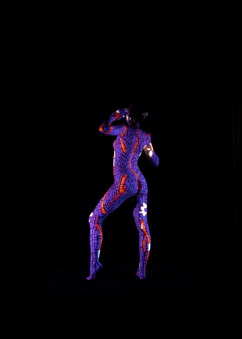 Person standing on tiptoes with body painted in a glowing pattern of purple, red, and white lines and shapes on a black background
